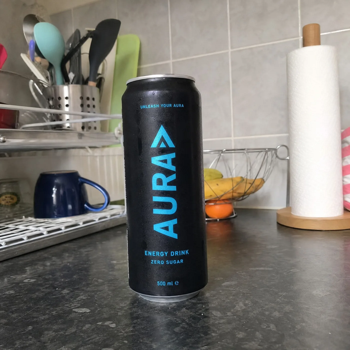 Onbewerkte telefoonfoto van een blikje energydrink op een keukenaanrecht, met afwasrek en fruitschaal op de achtergrond.